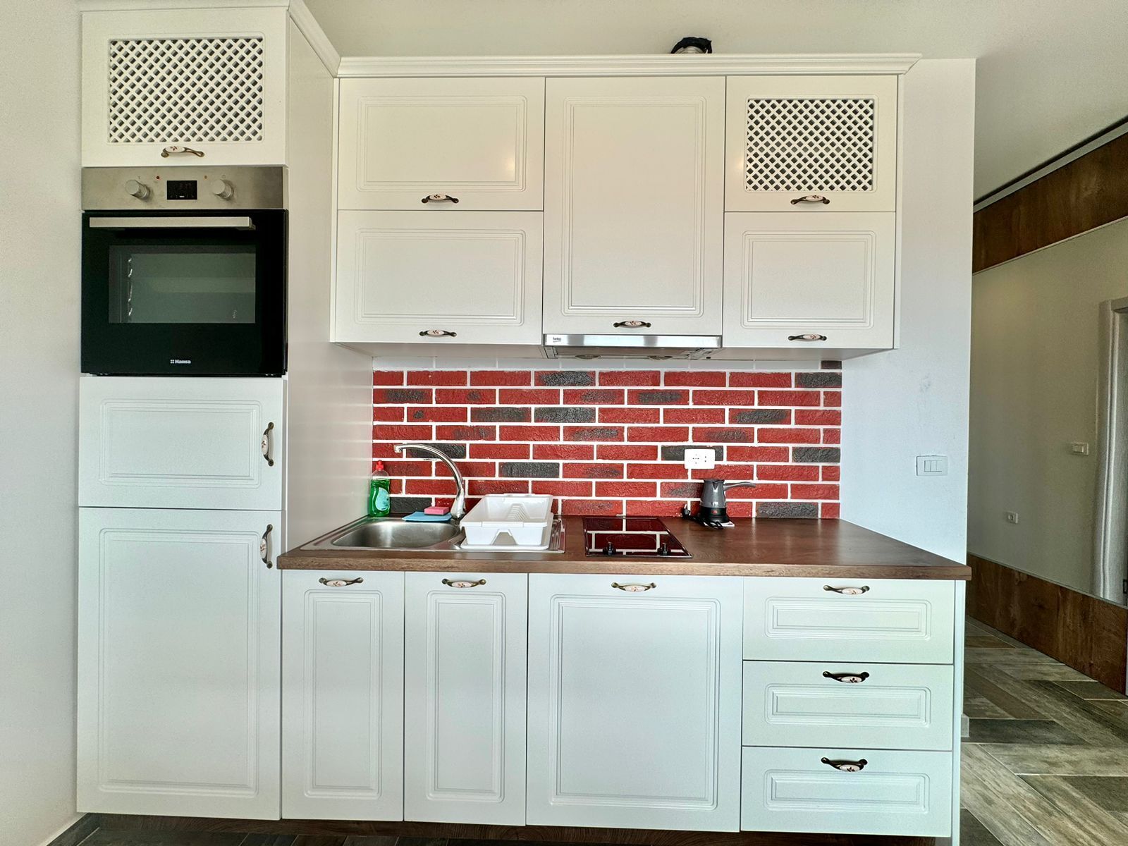 A compact kitchen with white cabinetry, a red brick backsplash, a built-in oven, and a sink with modern faucets.