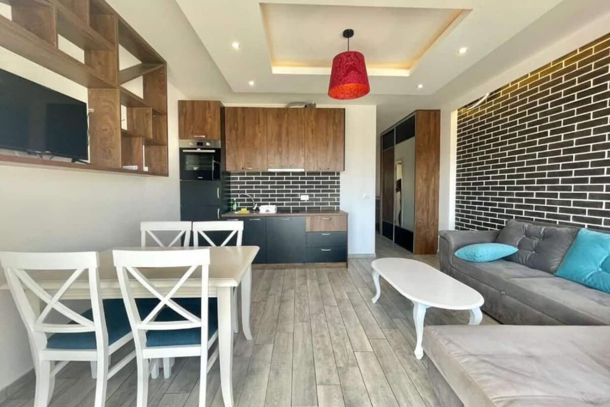 Una sala moderna con un sofá gris, sillas blancas, una mesa de centro blanca y una cocina con armarios de madera y salpicadero de ladrillo.