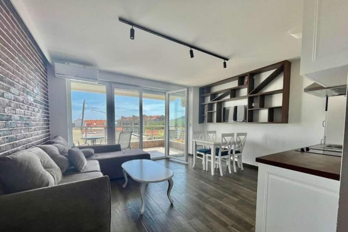 Una sala de estar moderna con un sofá, una mesa de centro y un comedor con sillas. Hay una cocina con una encimera y estanterías. La habitación tiene ventanas amplias y vistas al exterior.