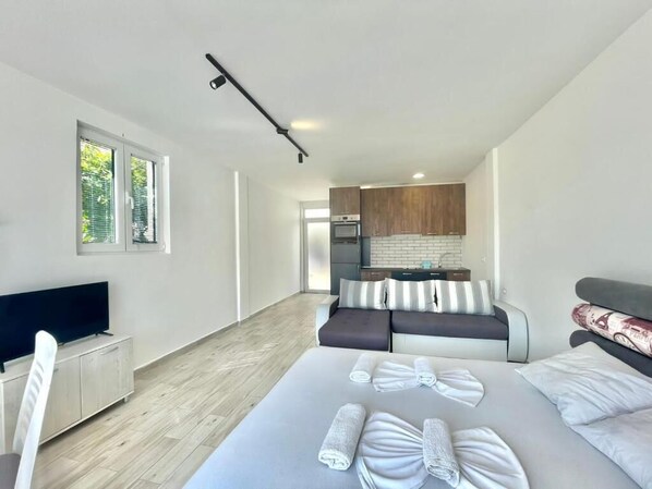 A modern hotel room with a large bed, a sofa, a TV, and a kitchenette.