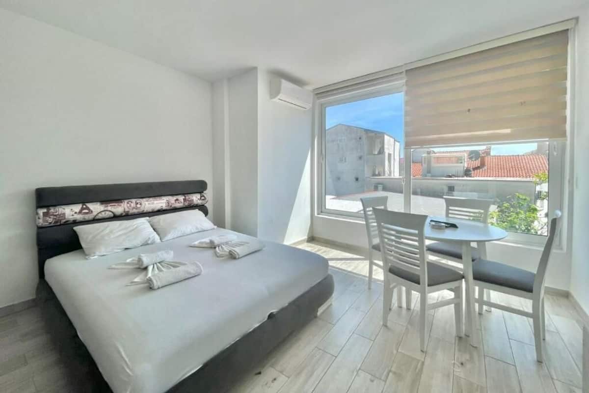 Habitación de hotel moderna con cama, una pequeña mesa con sillas y un gran ventanal con vistas a la ciudad.