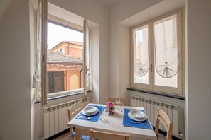 Uma área de refeições com uma mesa posta para dois, duas janelas com cortinas, um radiador abaixo das janelas e vista para edifícios lá fora.