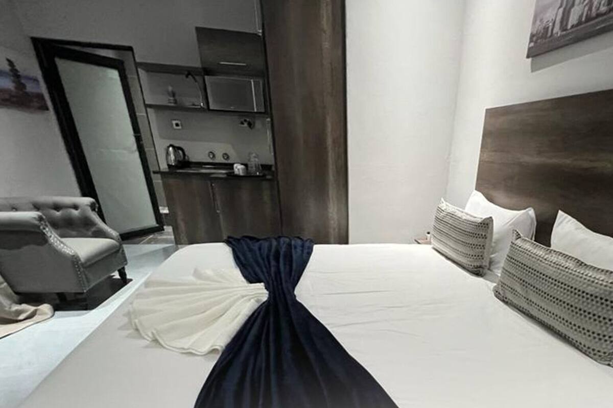 A hotel room with a neatly made bed, a chair, a kitchenette, and a large mirror.
