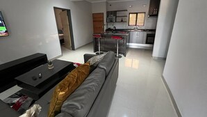 A modern living room with a grey sofa, a black coffee table, and a TV mounted on the wall. A kitchen with stainless steel appliances and a window is visible in the background. - Boalotswe Inn (Maun)