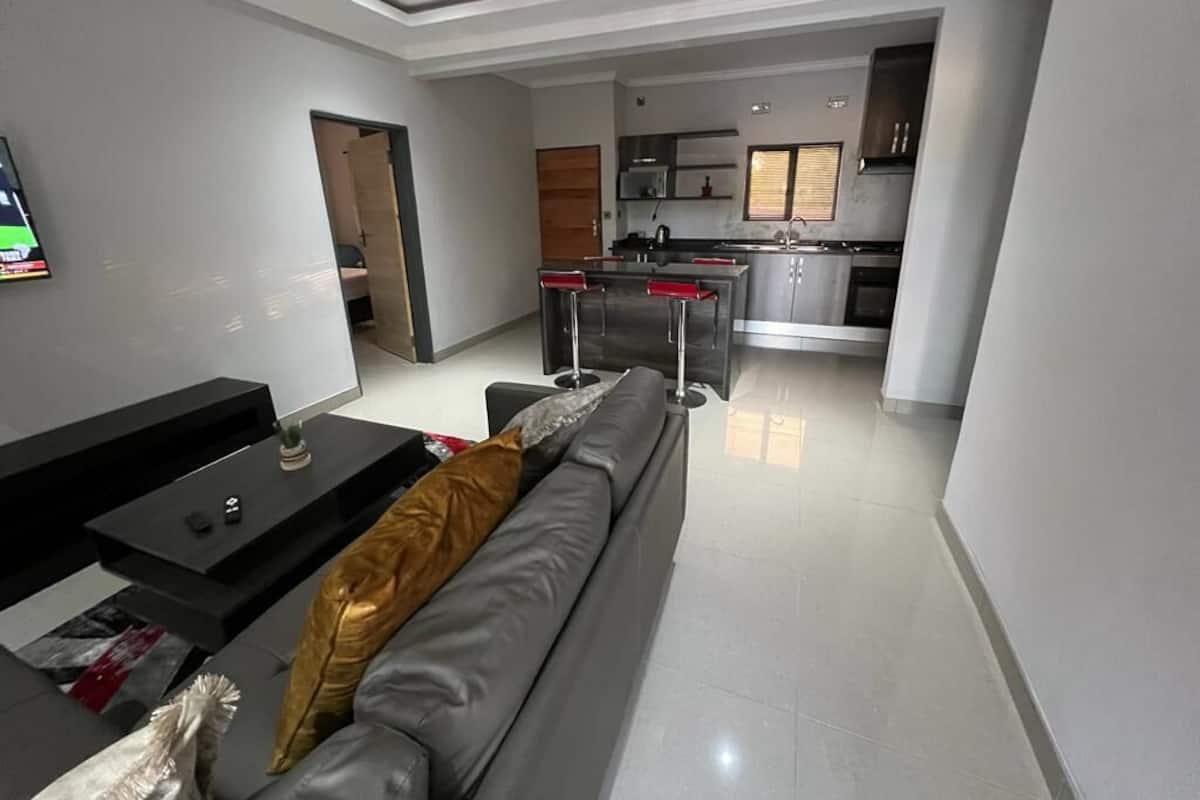 A modern living room with a grey sofa, a black coffee table, and a TV mounted on the wall. A kitchen with stainless steel appliances and a window is visible in the background.