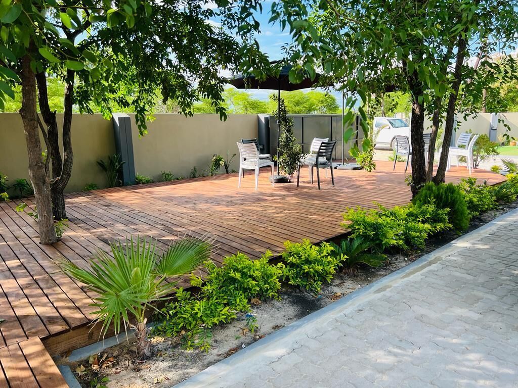 Um pátio externo com um deck de madeira, duas cadeiras brancas e uma pequena palmeira.