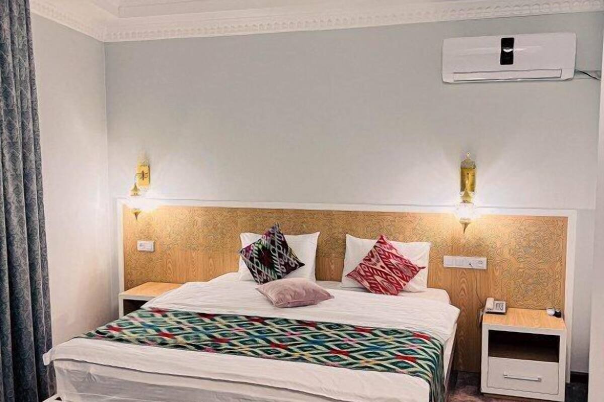 A hotel room with a large bed, a nightstand, an air conditioning unit, and a wall-mounted lamp.
