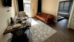 A dining area with a table set for four, a sofa, a TV mounted on the wall, and a window with blinds. - CAPPADOCIA SUIT RESIDENCE (Nevsehir)