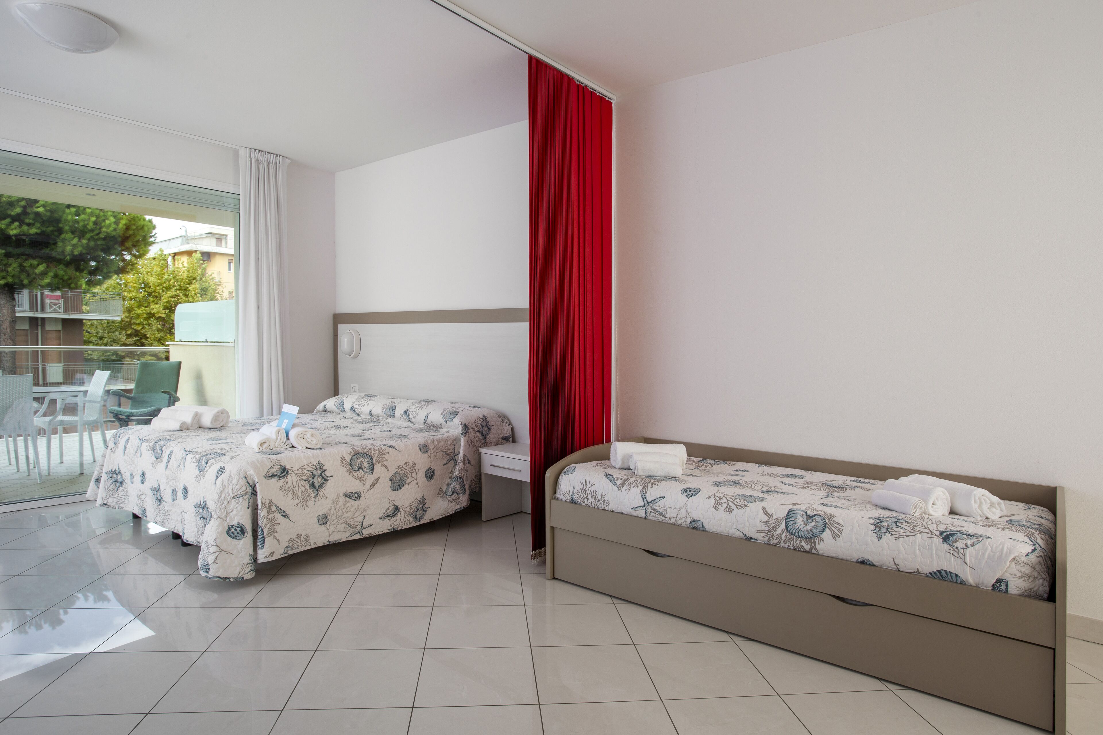 A hotel room with two beds, a red curtain, a balcony with a table and chairs, and a view of buildings and trees.