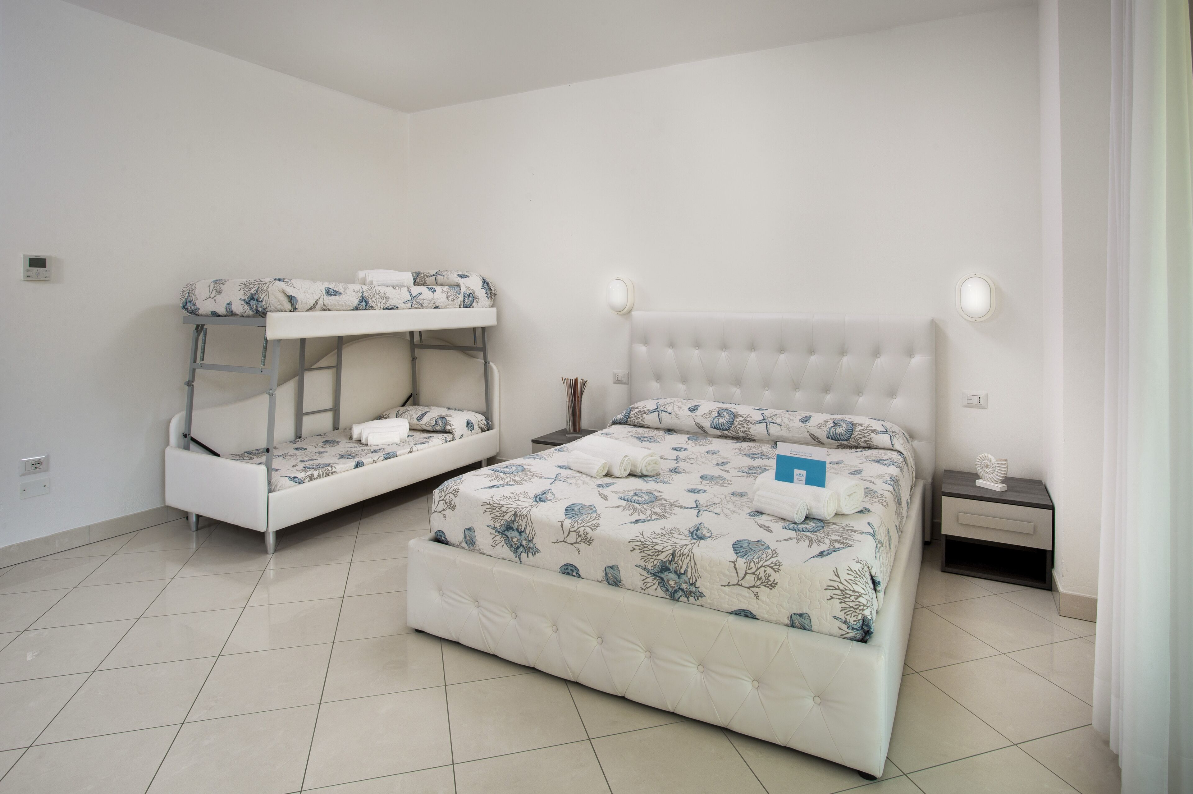 Una habitación con una cama litera y una cama individual, ambas con ropa de cama floral. Hay una mesita de noche con una lámpara y un pequeño objeto decorativo.