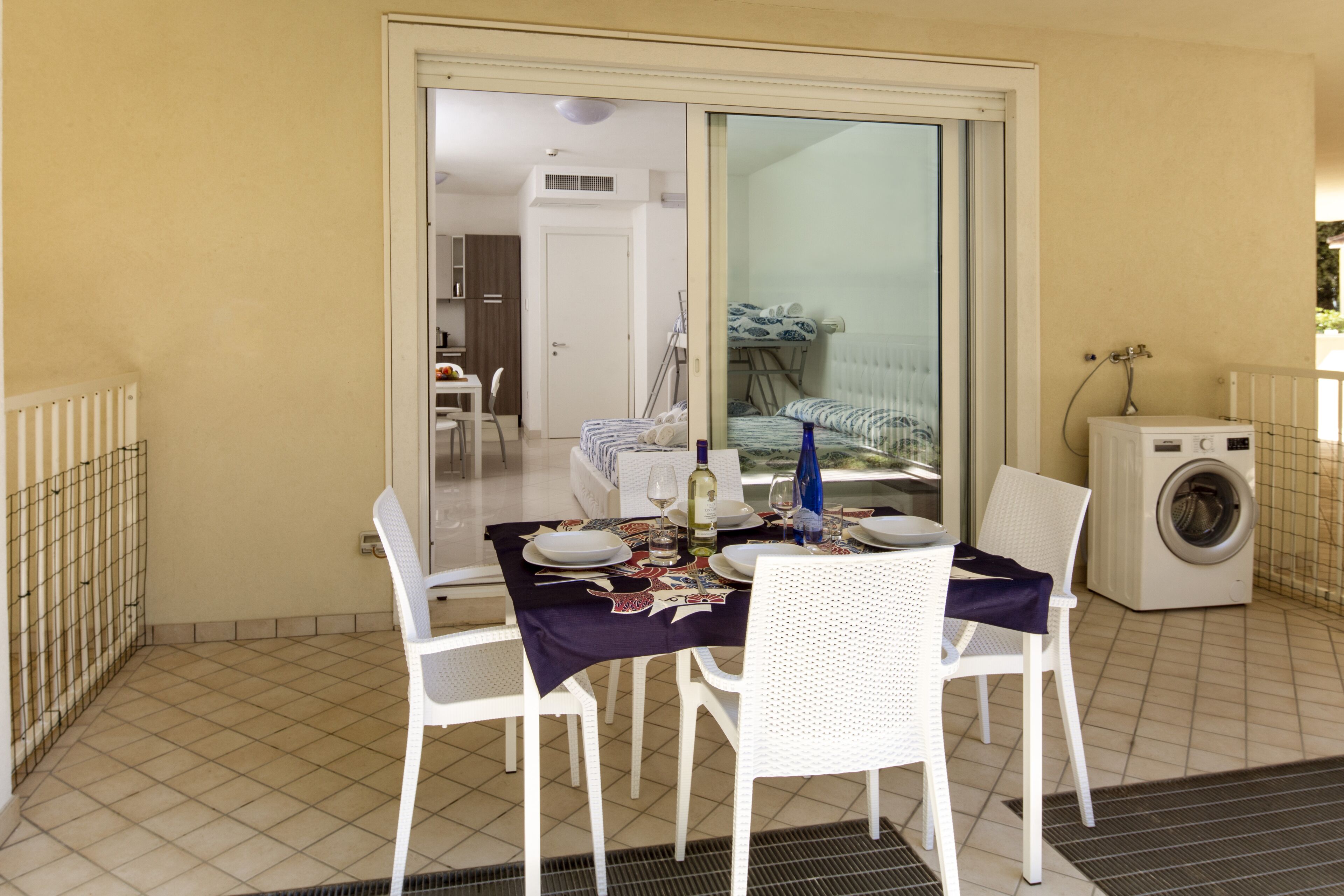 A balcony with a dining table set for two, white chairs, a washing machine, and a sliding glass door leading to a bedroom with a bed and a radiator.