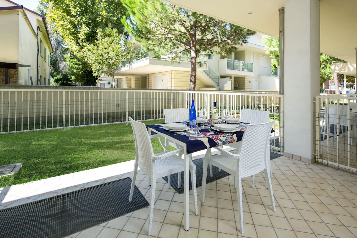 A balcony with a table set for four, surrounded by white wicker chairs, overlooking a grassy area and a building with balconies.