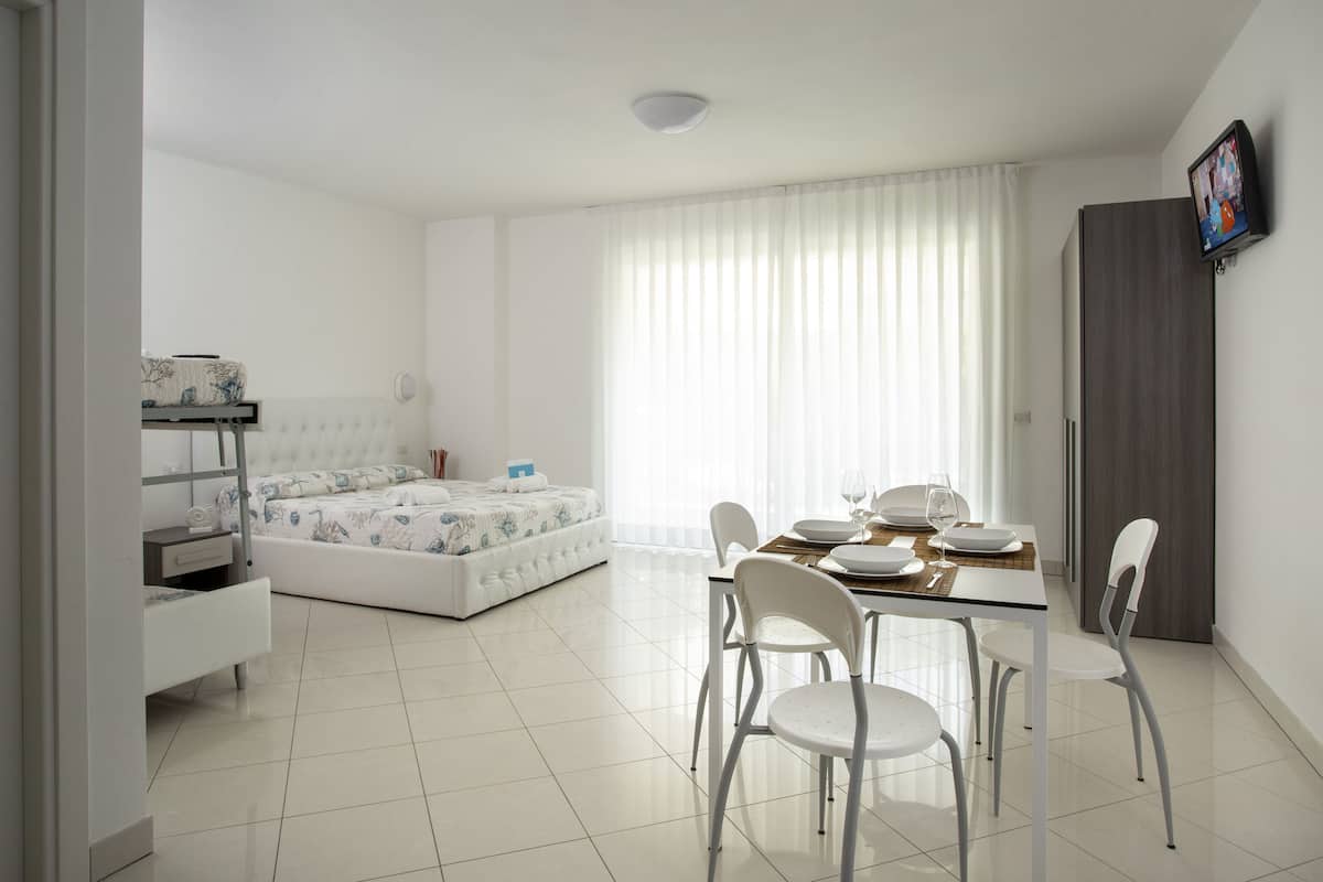 A modern, well-lit room with a bed, a dining table set for two, and a TV mounted on the wall.