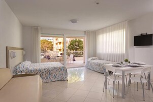 A hotel room with two beds, a dining table, chairs, a TV, and a balcony with a view of buildings and trees.
