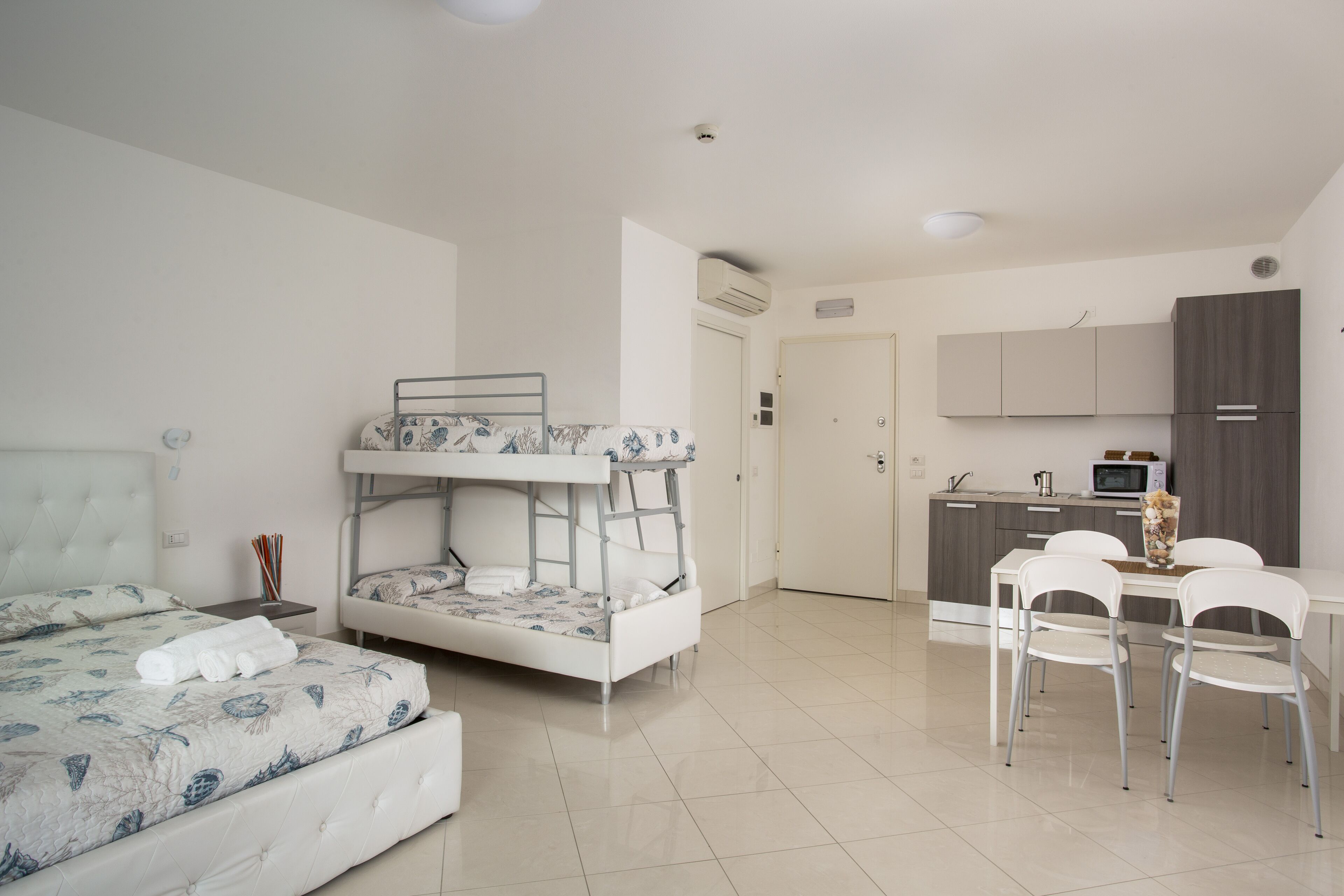 A bedroom with a bunk bed, a separate bed, a dining area with chairs, and a kitchenette.