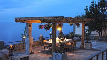 Un espacio de comedor al aire libre con una mesa dispuesta para dos personas, bajo una pérgola de madera con cestas de flores colgantes.