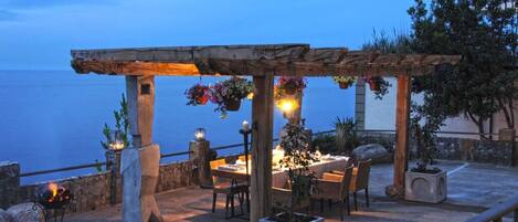Un espacio de comedor al aire libre con una mesa dispuesta para dos personas, bajo una pérgola de madera con cestas de flores colgantes.