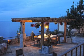 An outdoor dining area with a table set for two, under a wooden pergola with hanging flower baskets.