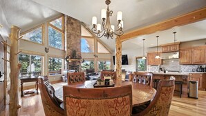 Un comedor espacioso con una gran mesa de madera y sillas estampadas, una araña y ventanas amplias que ofrecen vistas del exterior.