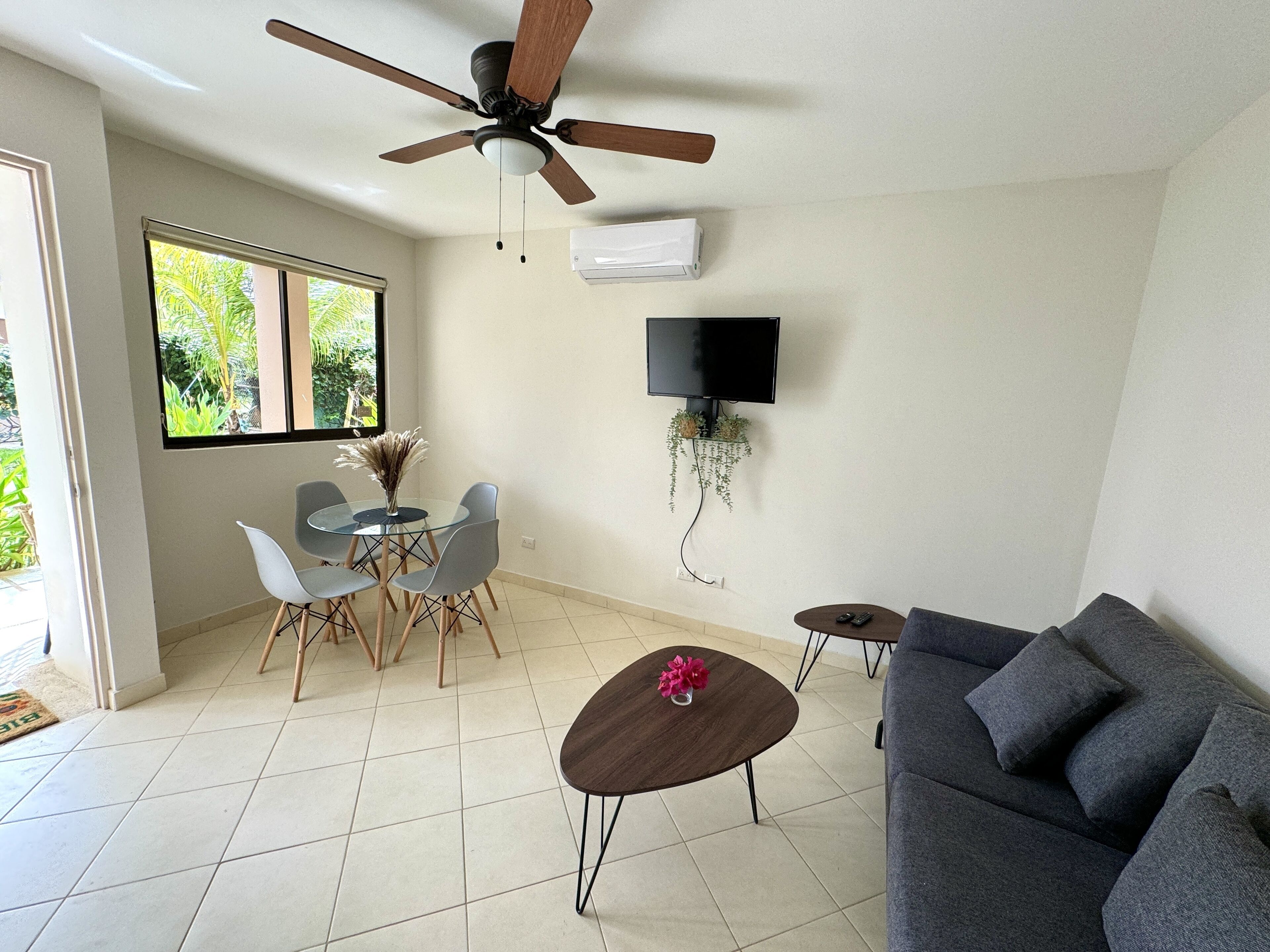 Un salon doté d’un ventilateur de plafond, d’un téléviseur fixé au mur, d’une table à manger avec des chaises, d’un canapé et d’une petite table basse.