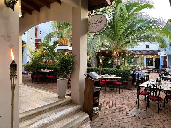 An outdoor dining area with tables and chairs, a sign for 'La Dolce Vita International Restaurant & Pizzeria,' and a large potted plant.