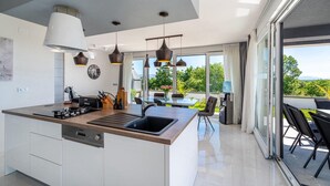 Una cocina moderna con una isla central, un comedor con mesa de vidrio y vistas a la vegetación a través de amplias ventanas.