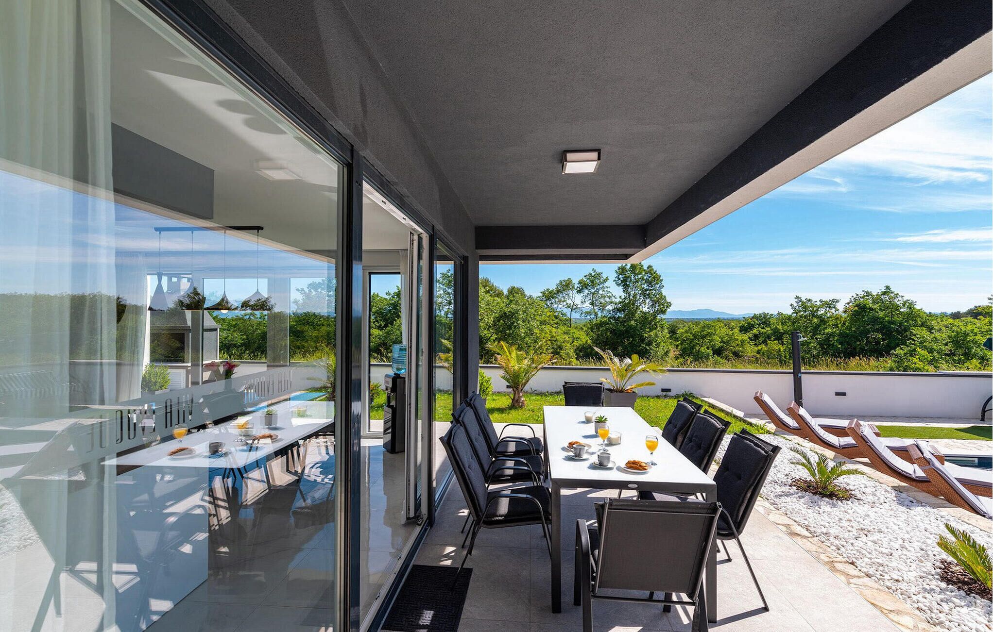 A modern outdoor dining area with glass walls, a pool, and a view of greenery and mountains.