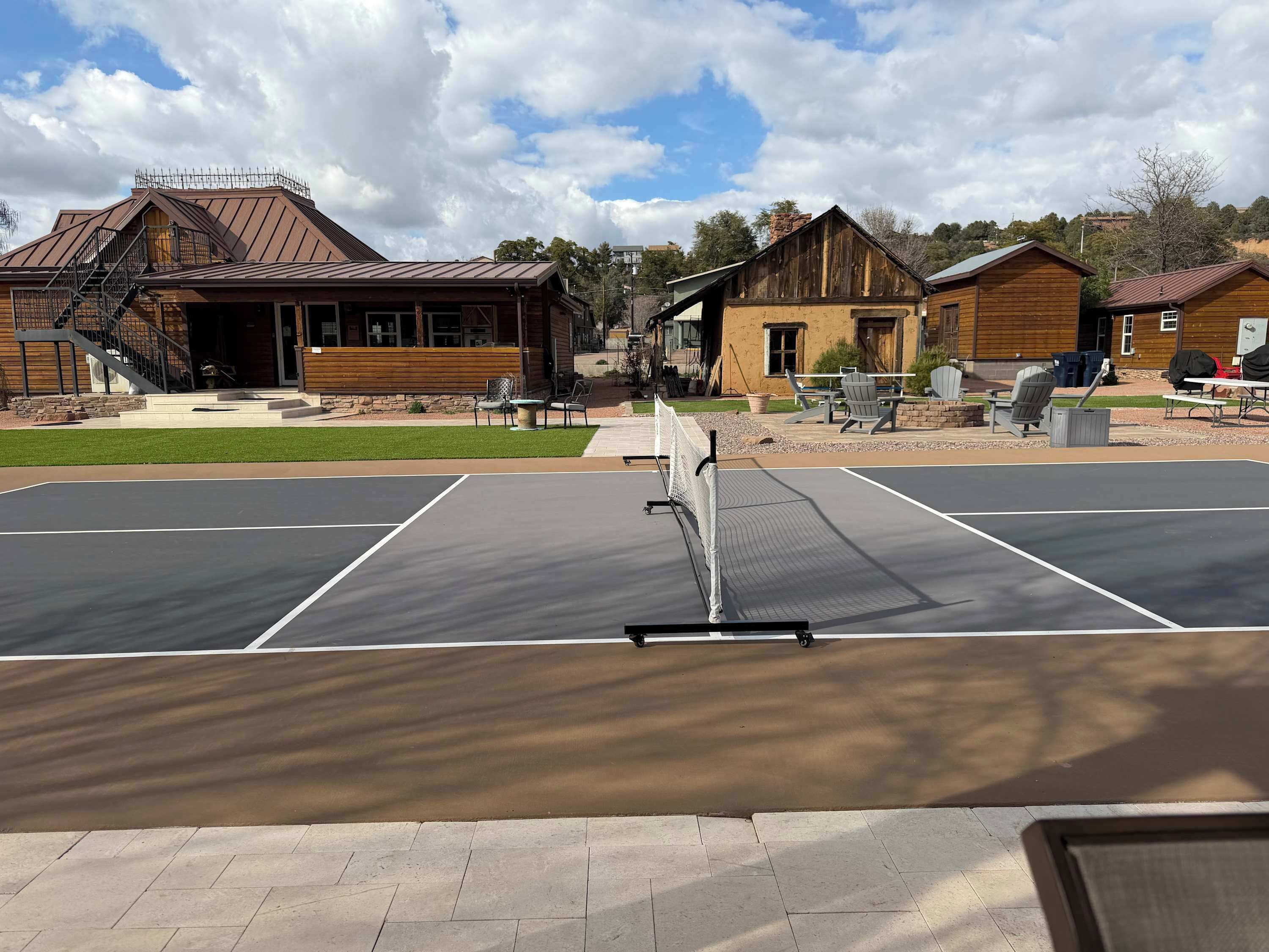 Ein Tennisplatz mit Netz im Vordergrund, umgeben von Außenplätzen und Holzbauten im Hintergrund.