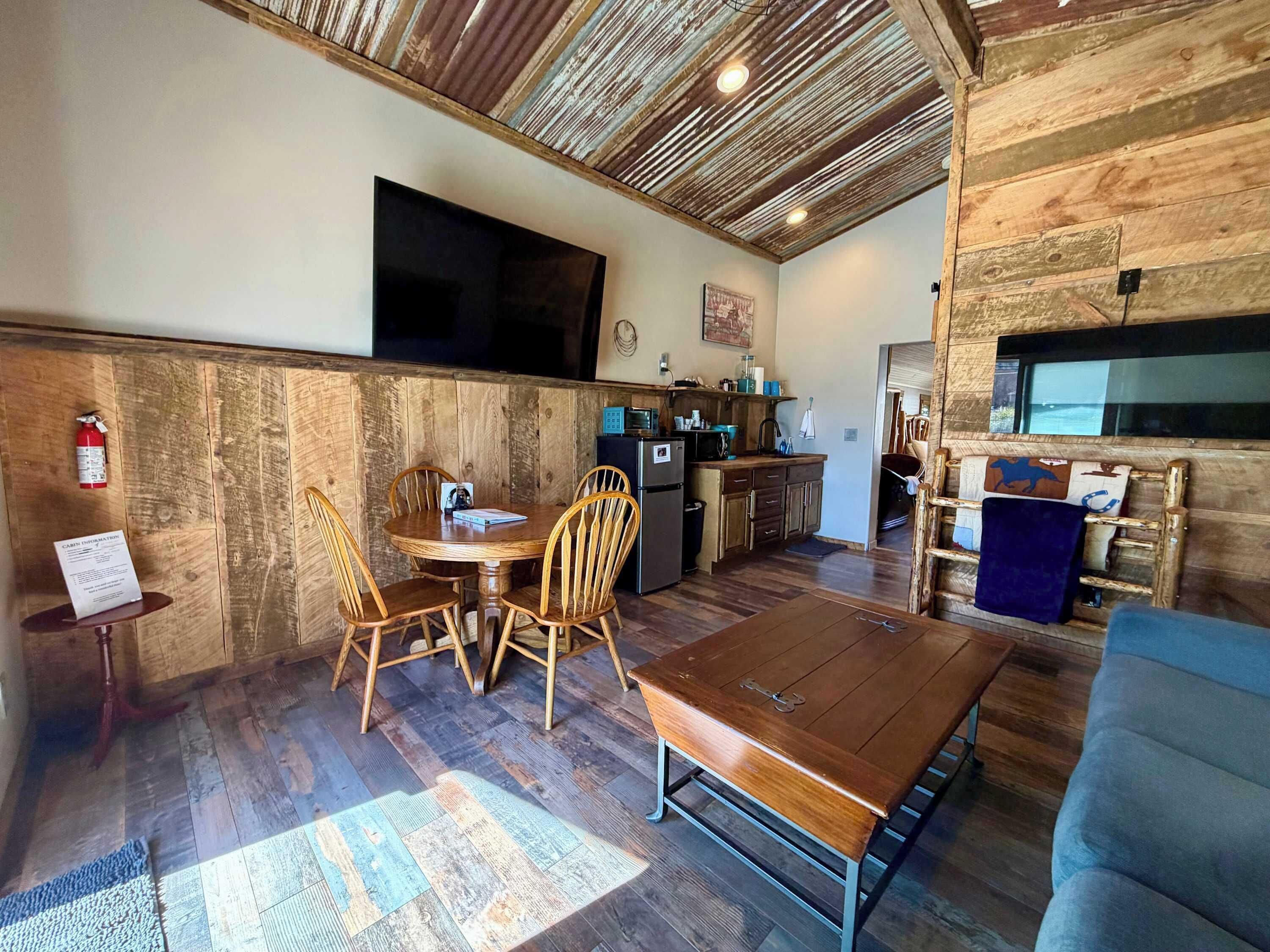 Una habitación acogedora con techo de madera, una mesa de comedor con sillas, un sofá y una zona de cocina con microondas y fregadero.