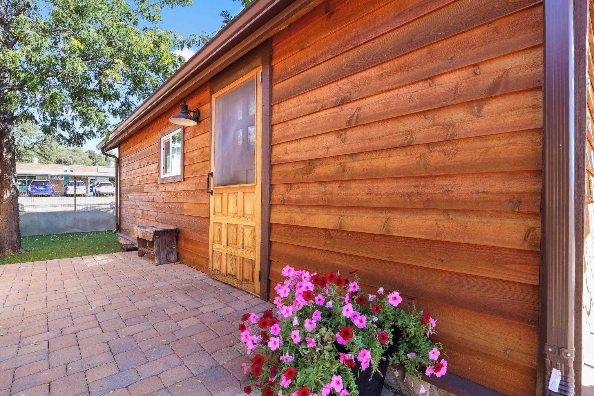 Una cabaña de madera con una puerta grande y una ventana, un camino de ladrillo y un macizo de flores con flores rosadas y rojas.