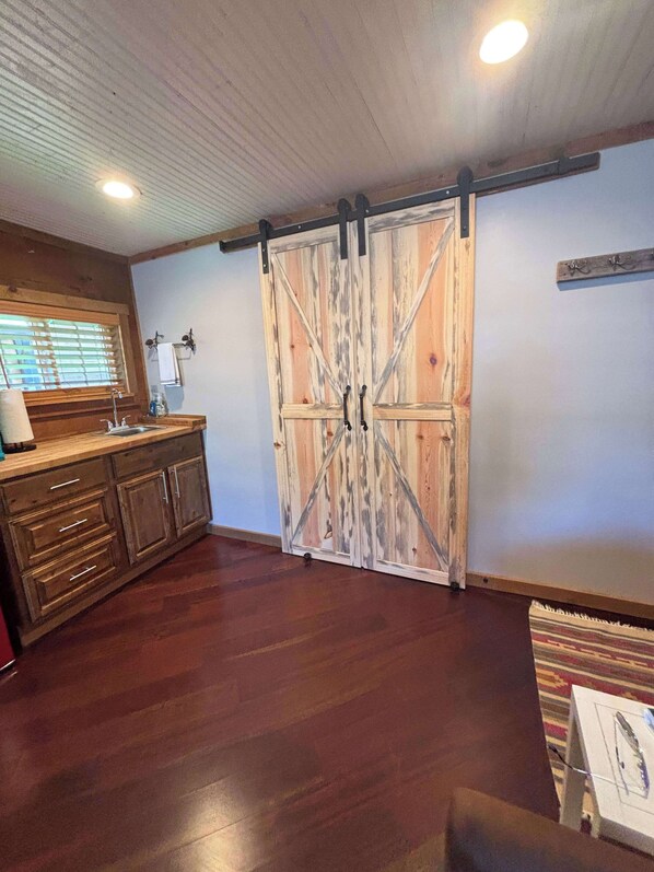 A rustic kitchen with wooden cabinets, a farmhouse-style sliding barn door, and a window with blinds. - Mini Cabin stay with pickleball court! (Payson)