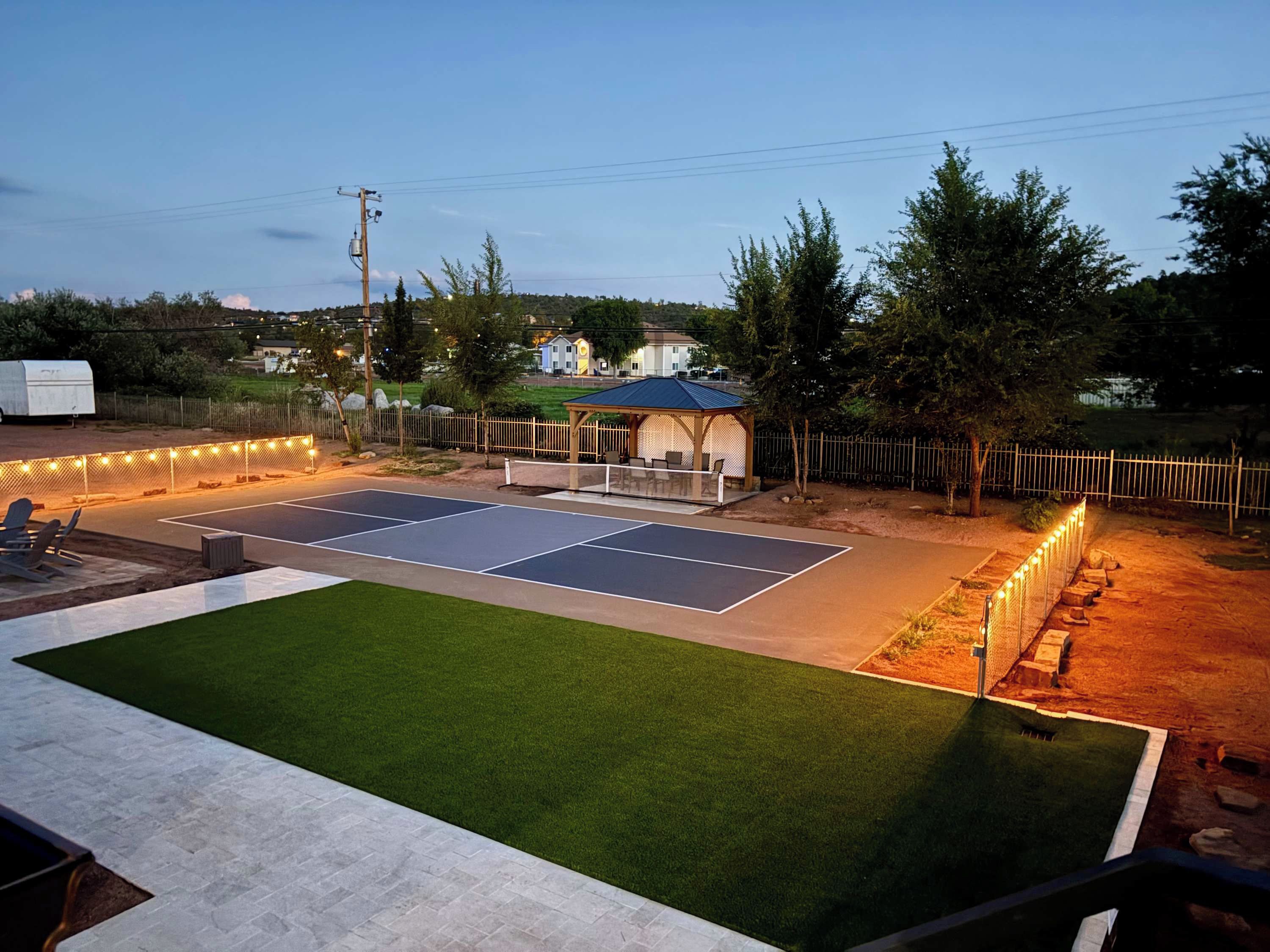 A tennis court with a covered seating area and a gazebo, surrounded by a fence and trees.