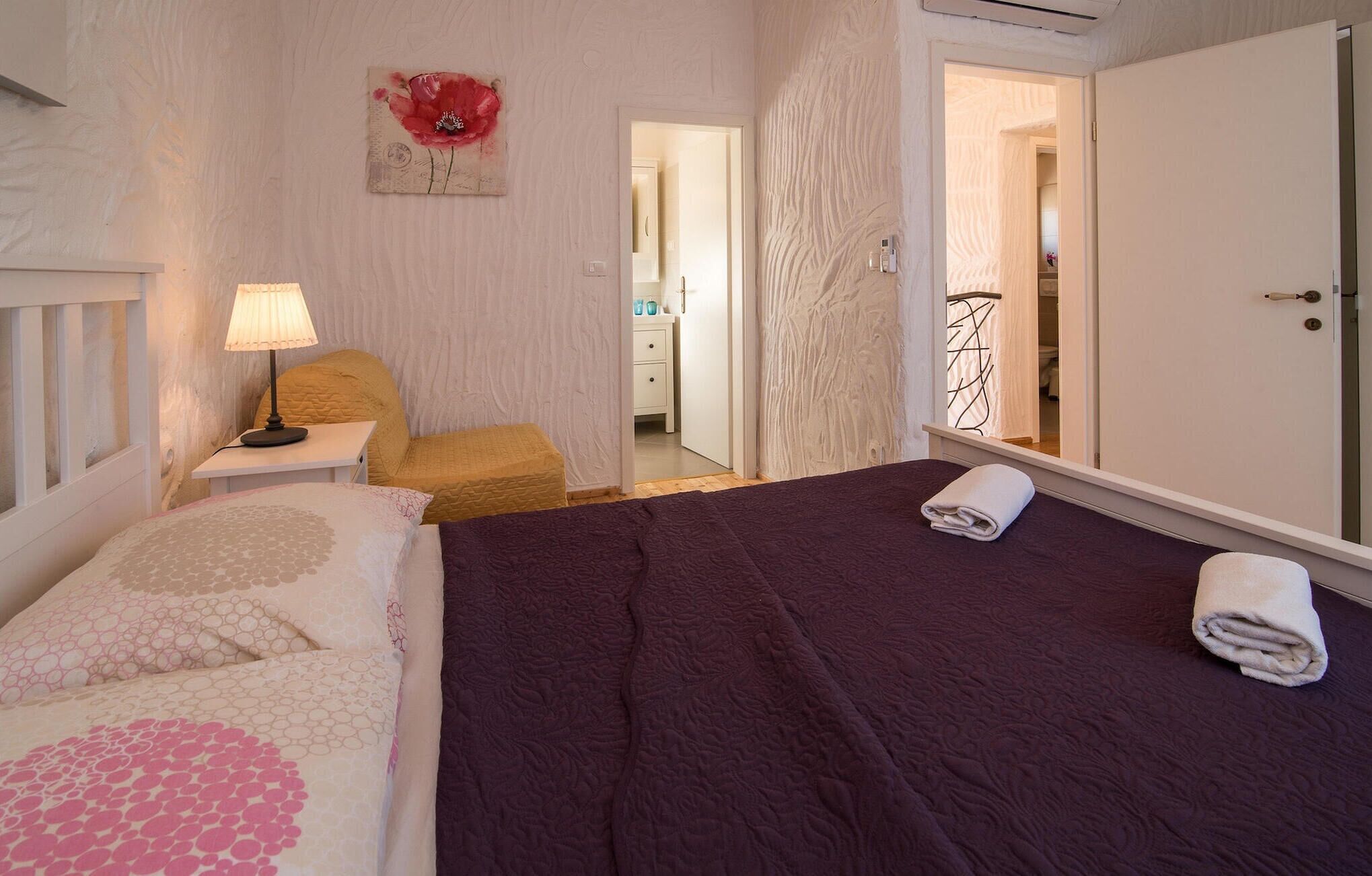 Un dormitorio con cama, mesita de noche, lámpara, silla y puerta que da a un baño.
