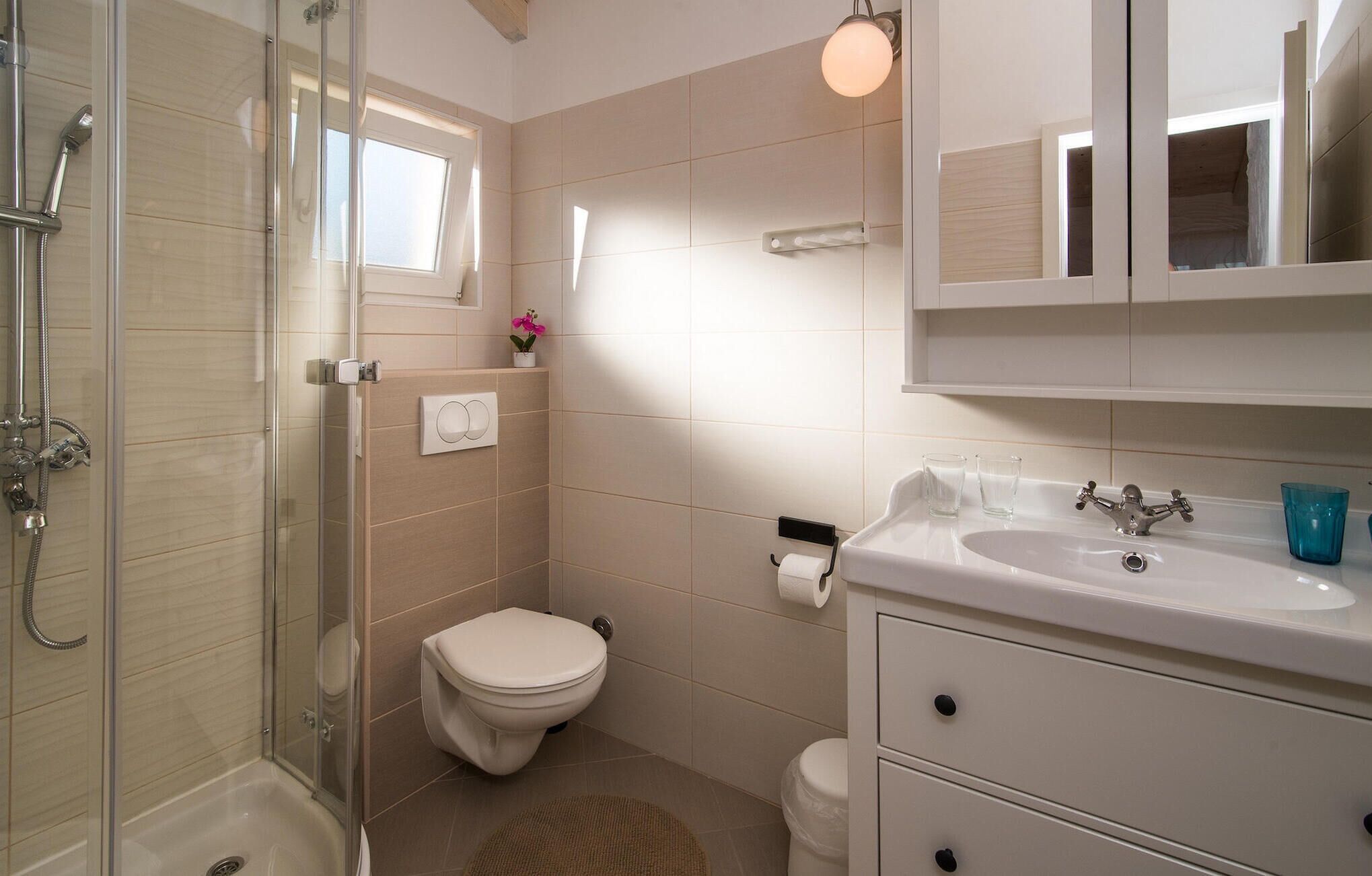 A bathroom with a glass-enclosed shower, a toilet, a sink with a mirror, and a small window.