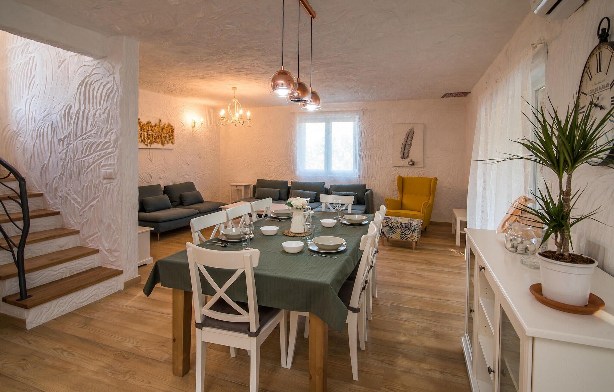 Uma sala de jantar com uma mesa redonda posta para seis pessoas, rodeada por cadeiras brancas. Há uma poltrona amarela e um sofá cinzento ao fundo.
