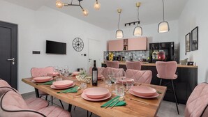 A modern dining area with pink chairs, a wooden table set for six, and a kitchen in the background.