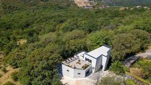A modern, single-story house surrounded by dense forest, with a flat roof and a small patio area.