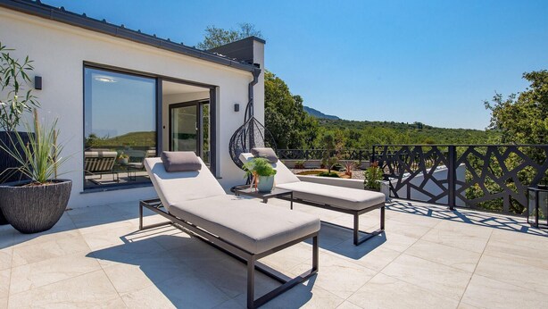 A modern outdoor seating area with two lounge chairs and a table, surrounded by potted plants and a view of a mountainous landscape.