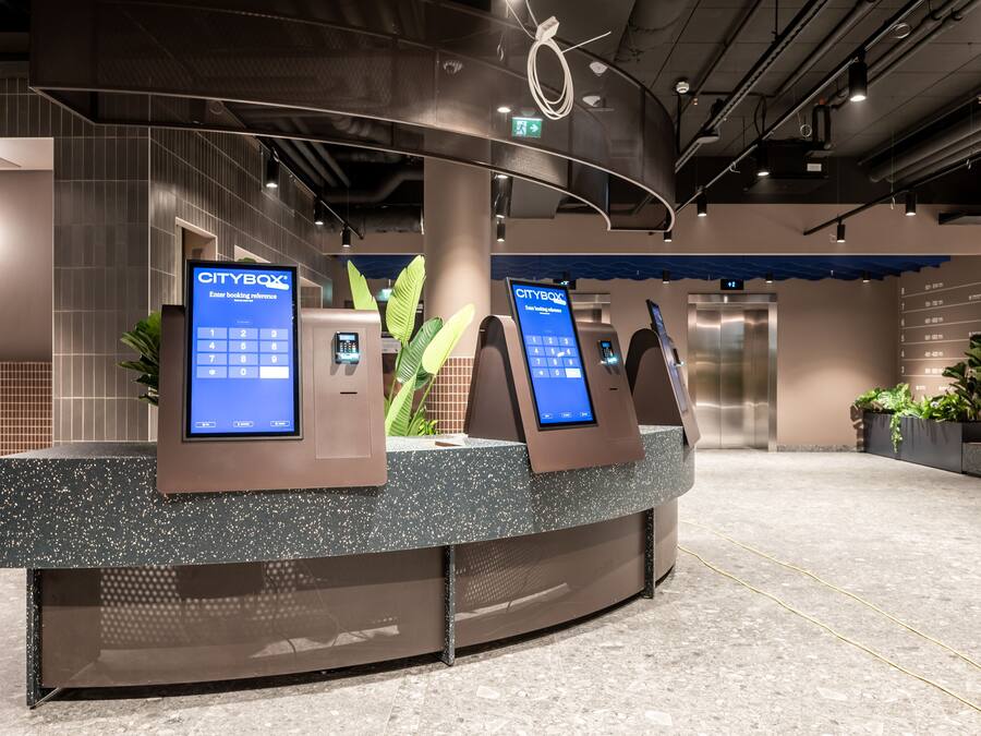 Un área de recepción moderna con un mostrador curvo, dos quioscos digitales y un ascensor al fondo.