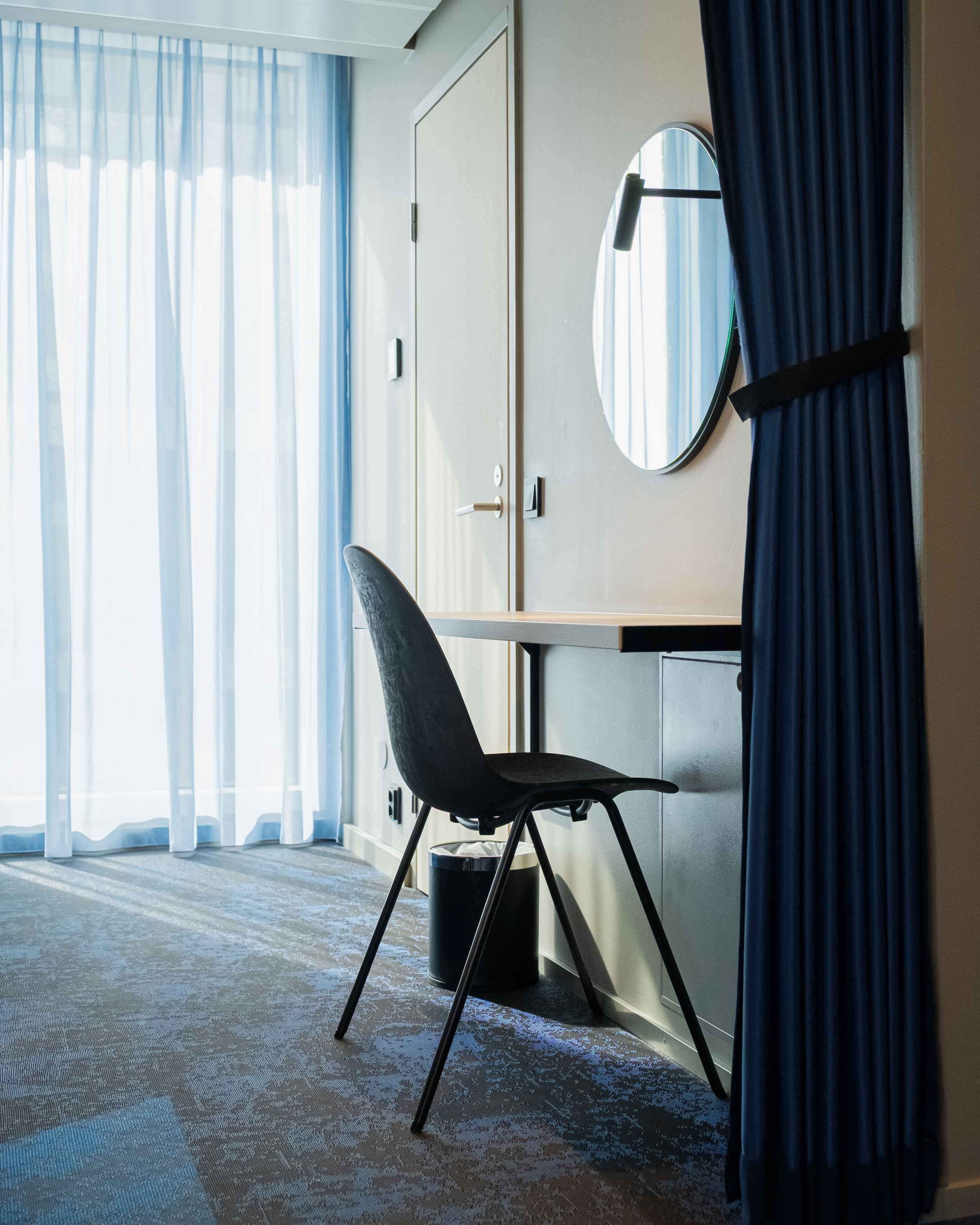 Una habitación de hotel con un ventanal grande, una silla, un escritorio, un espejo redondo y un armario.