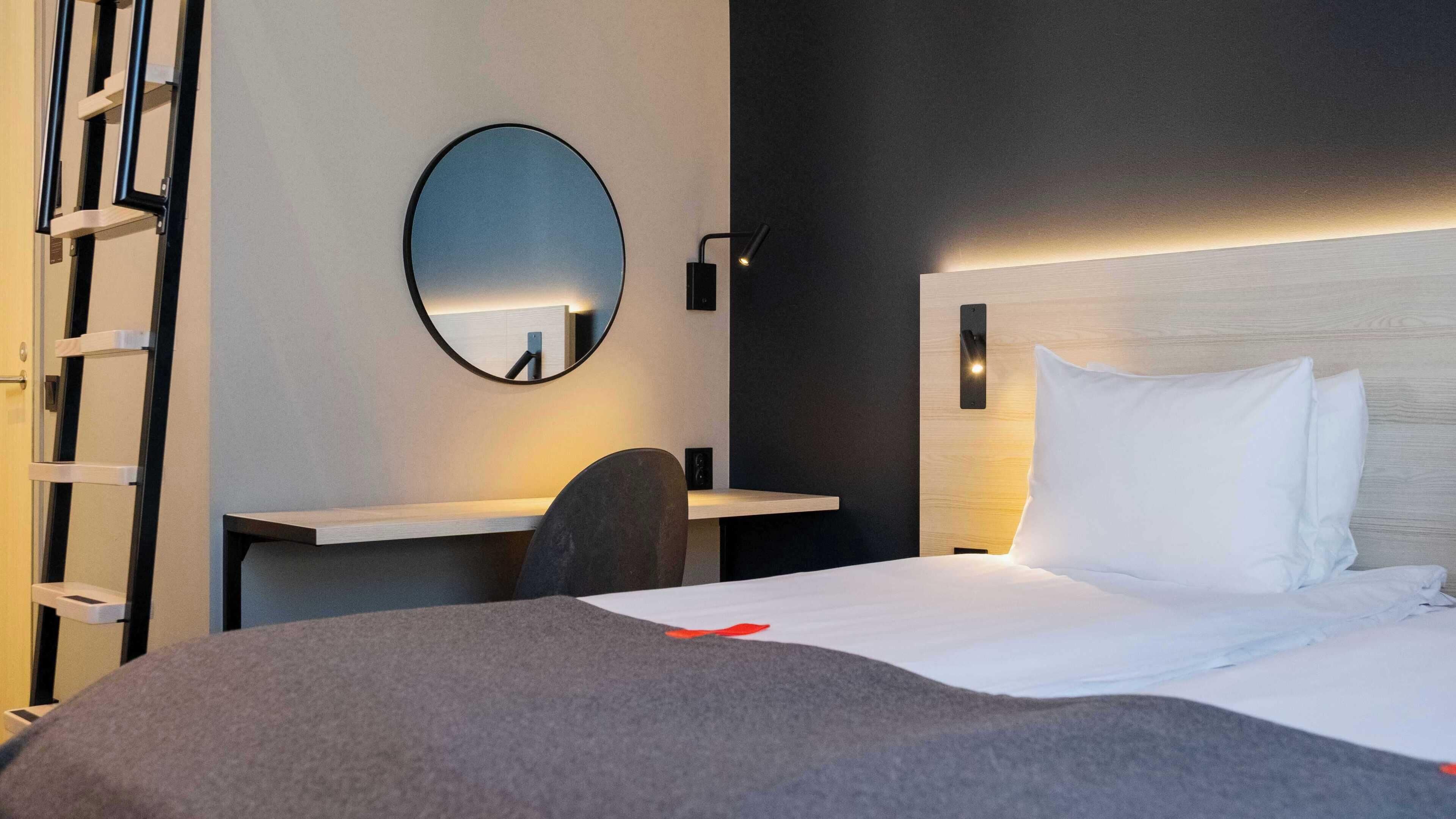 Habitación de hotel con cama, escritorio, silla, espejo y lámpara de pared.