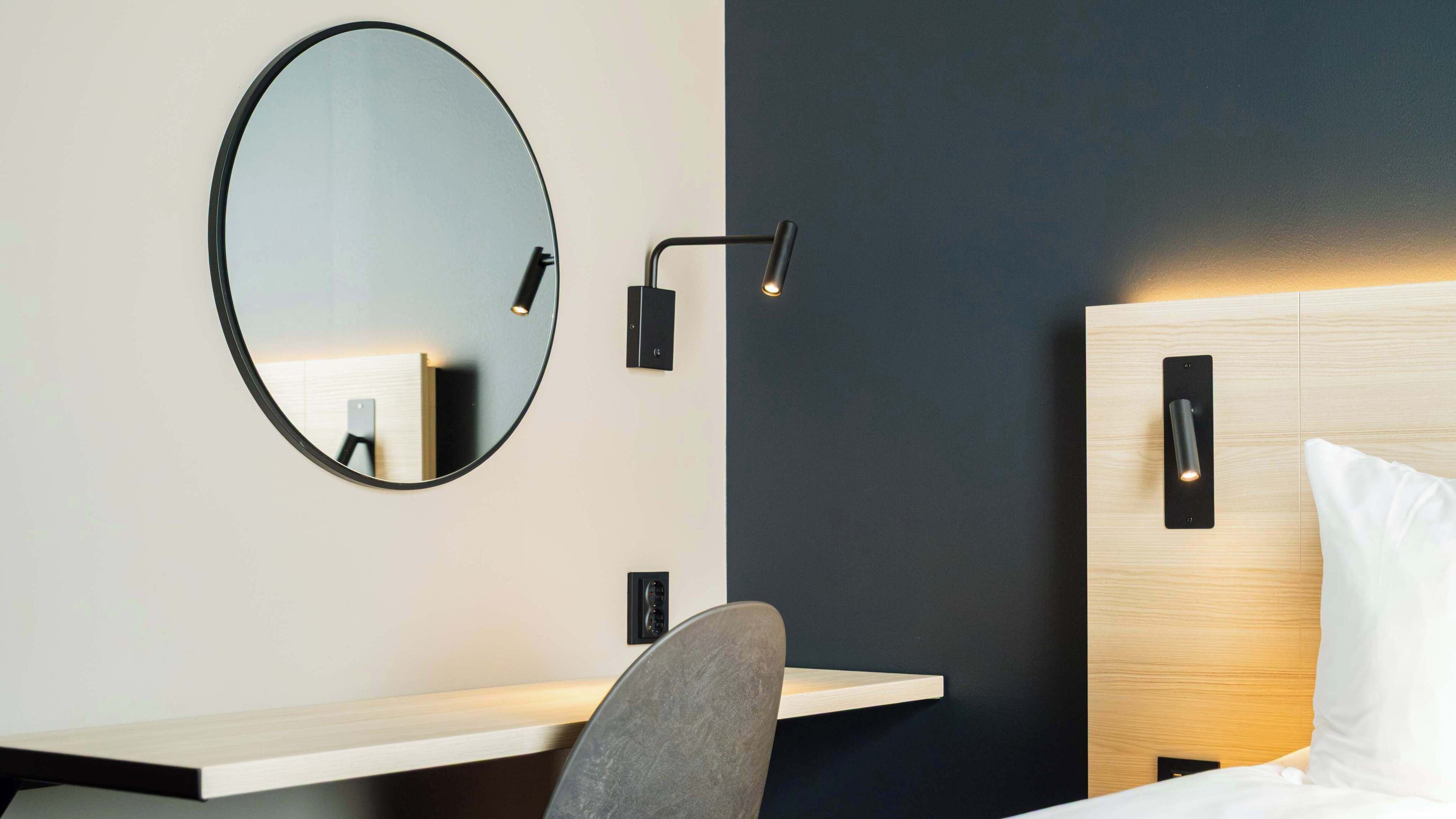 A hotel room with a round mirror, bedside table, chair, and a bed with a headboard.
