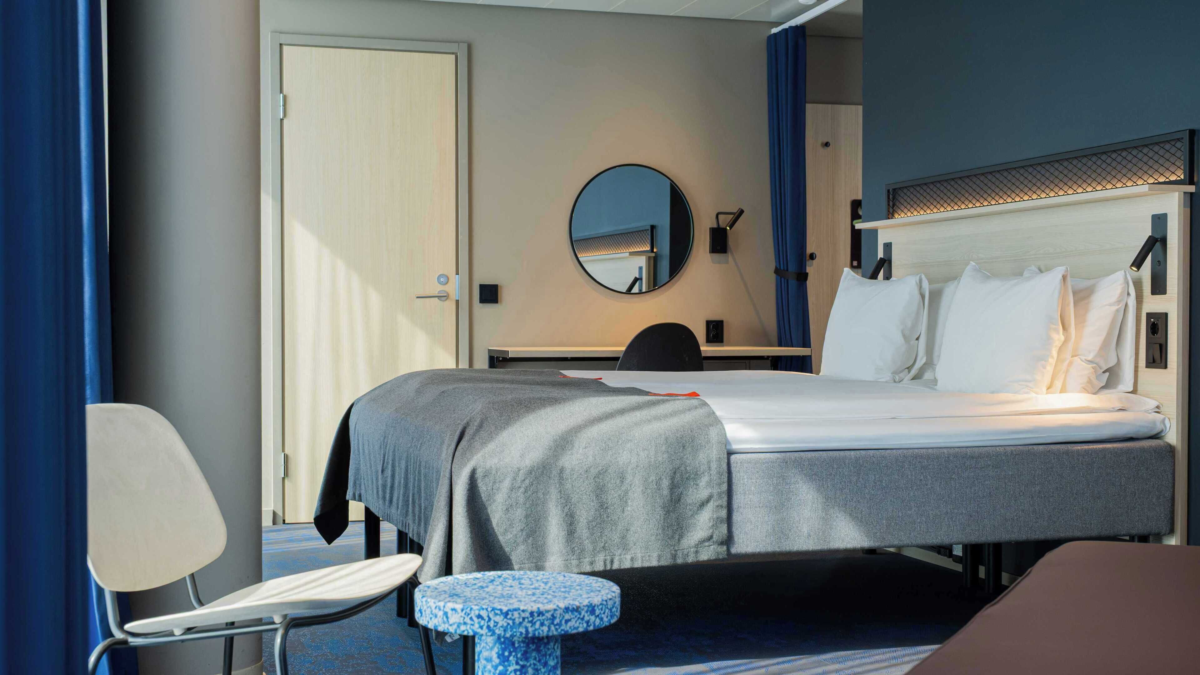 Quarto de hotel com cama, escrivaninha, cadeira, mesa redonda pequena e espelho.