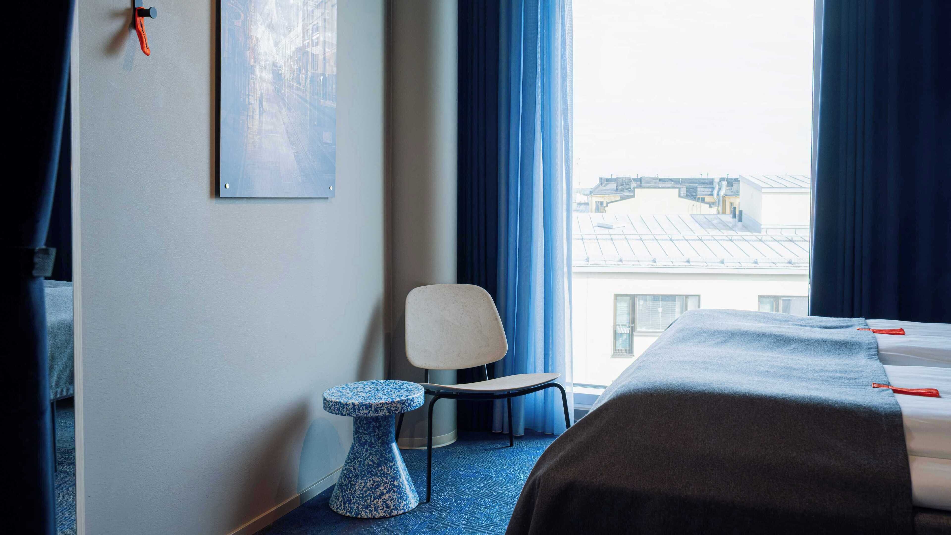 A hotel room with a bed, a chair, a small table, and a window with a view of buildings.