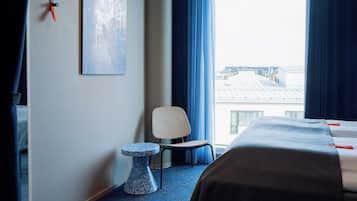A hotel room with a bed, a chair, a small table, and a window with a view of buildings.