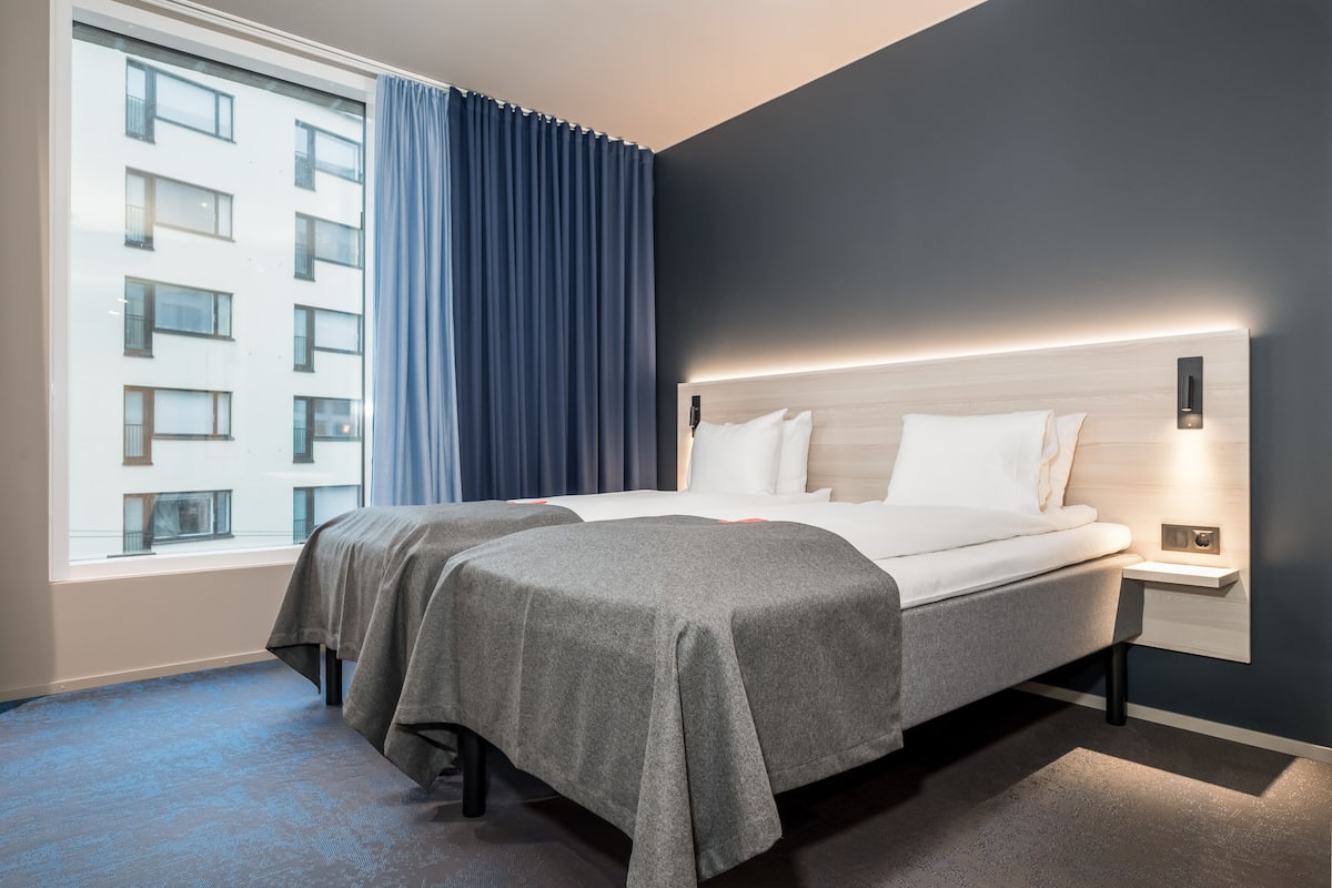 A modern hotel room with two beds, a large window with curtains, and a nightstand with a lamp.