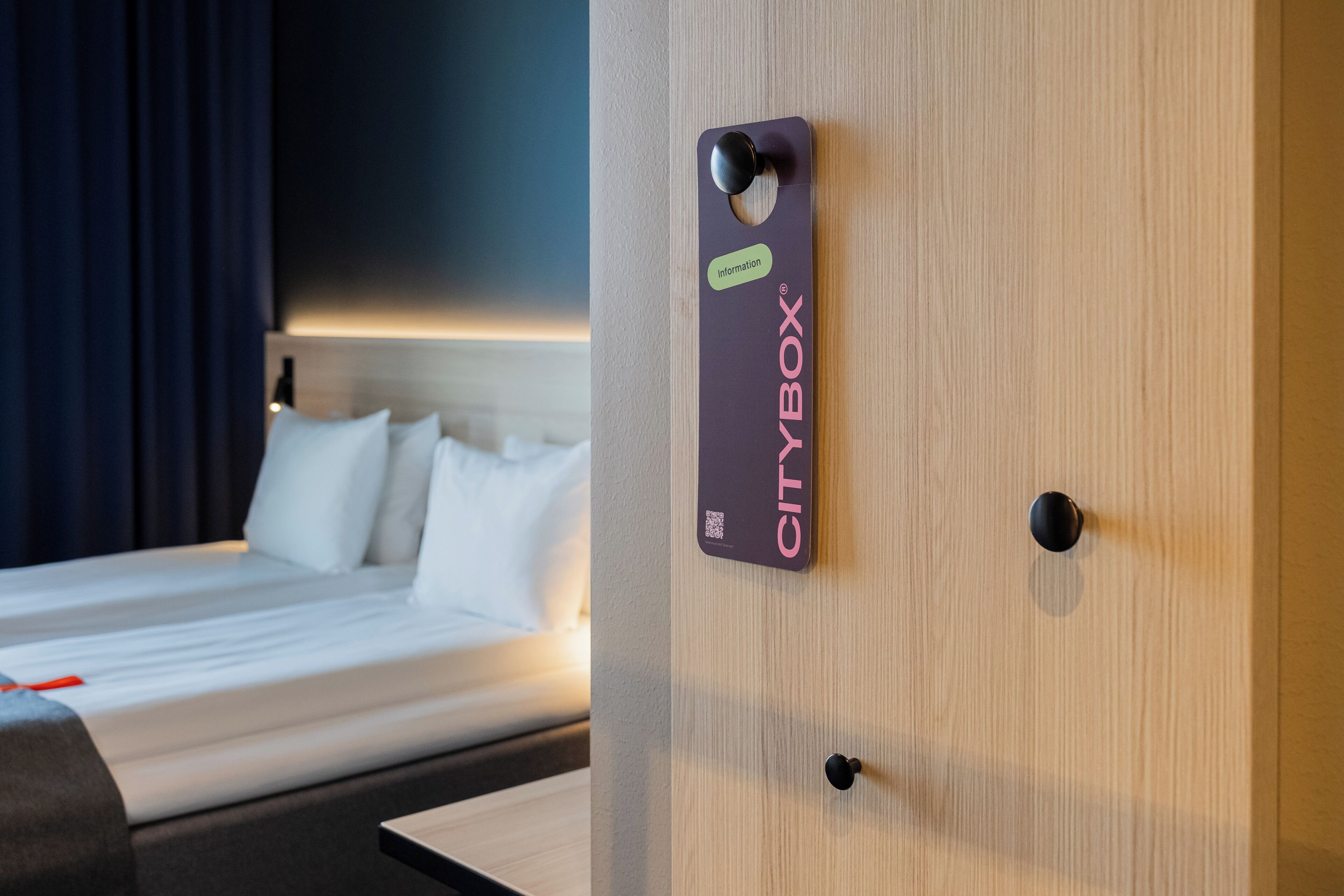 A CityBox® sign on a wooden door, a bed with white linens, and a dark curtain in a hotel room.