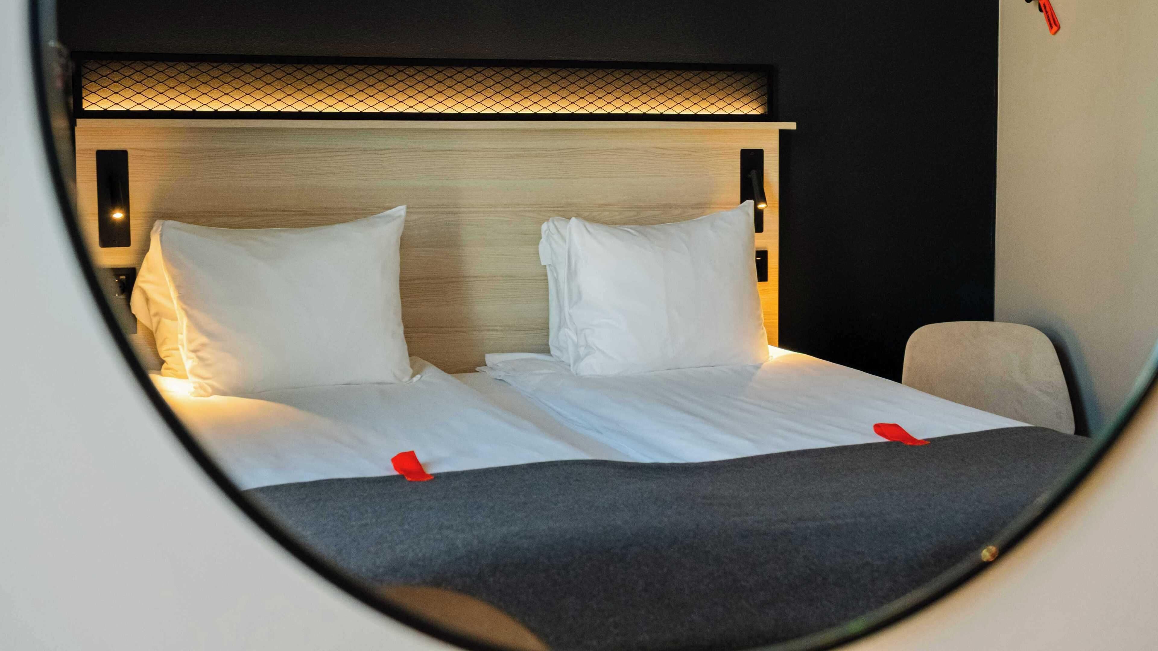 Un letto con lenzuola bianche e due cuscini, una testiera con pannello illuminato e una parziale vista di una sedia.