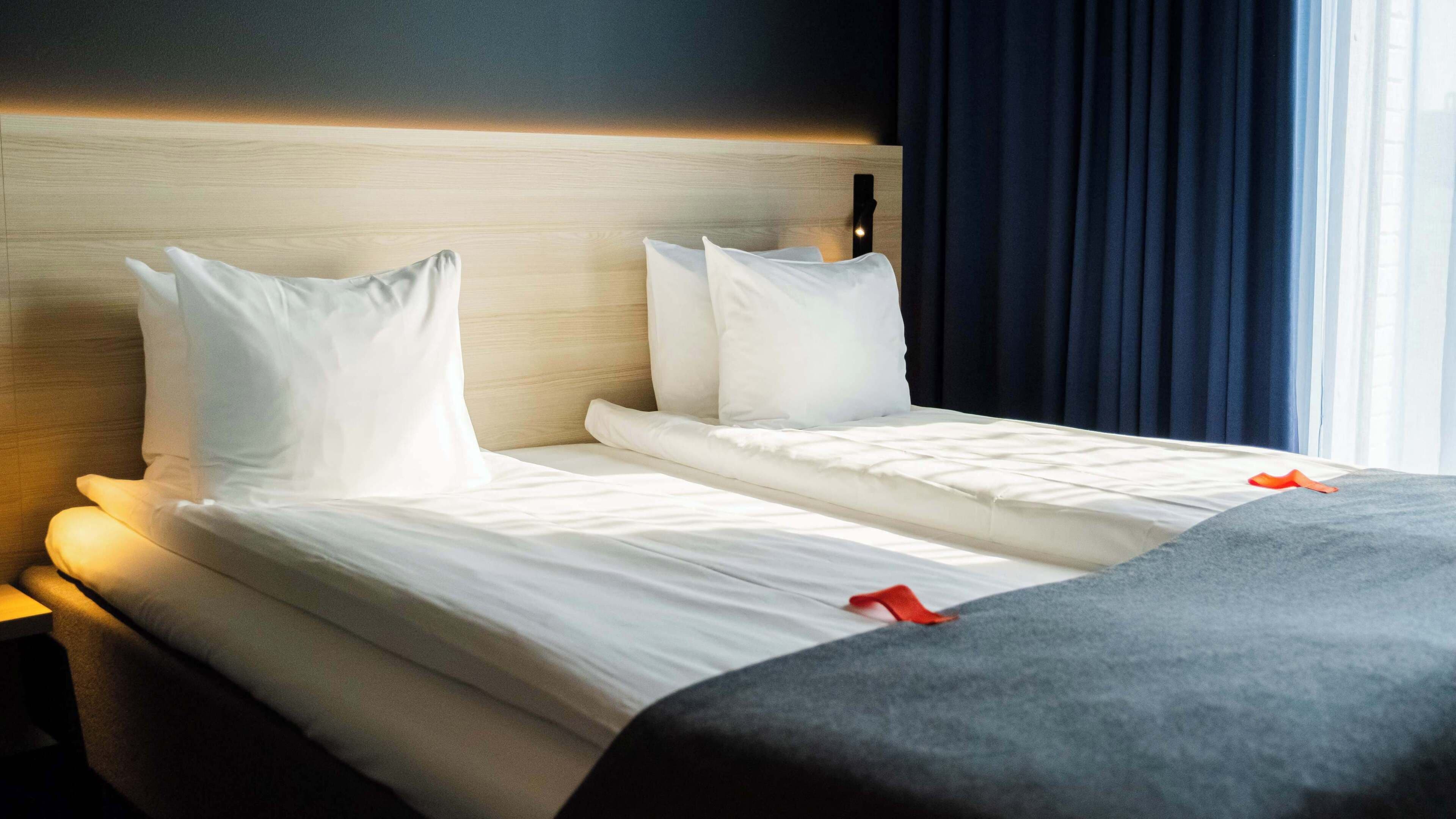 A double bed with white linens and a grey throw blanket, a wooden headboard, and a nightstand with a lamp.