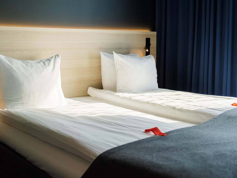 A double bed with white linens and a grey throw blanket, a wooden headboard, and a nightstand with a lamp.